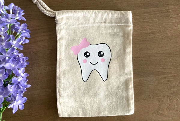 Small canvas tooth fairy bag with a smiling tooth design and pink bow on a wooden table