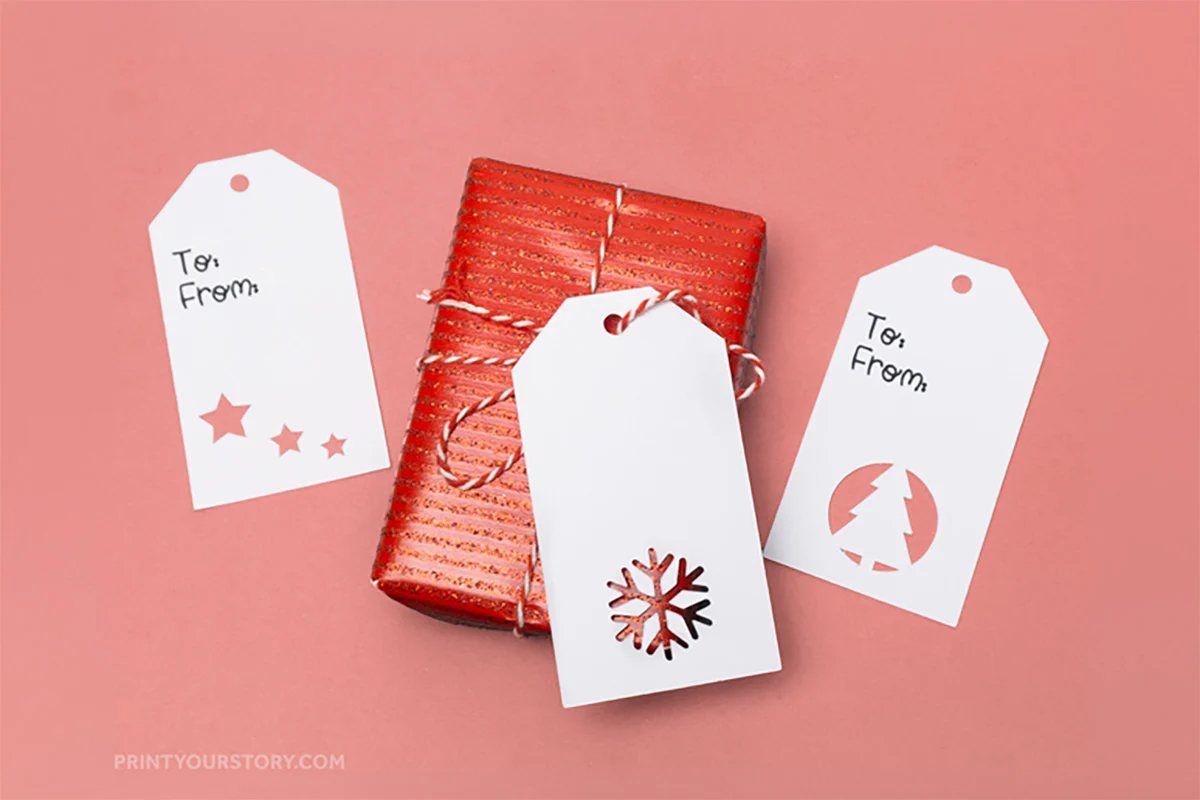 Three white Christmas gift tags with cutout star, snowflake, and tree designs around a red wrapped present