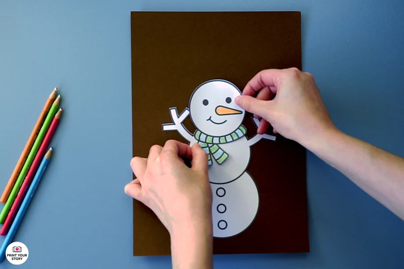 Hands pasting a paper snowman face onto a black sheet for a printable coloring craft
