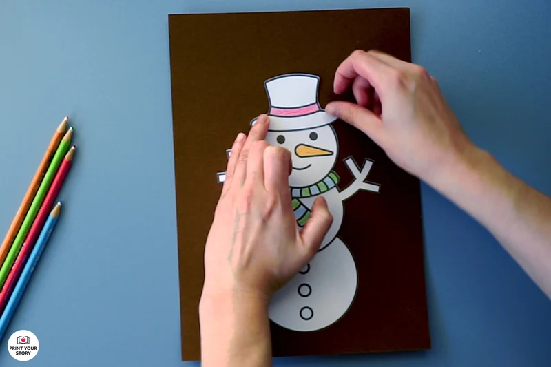 Hands pasting a paper hat onto a printable snowman coloring craft