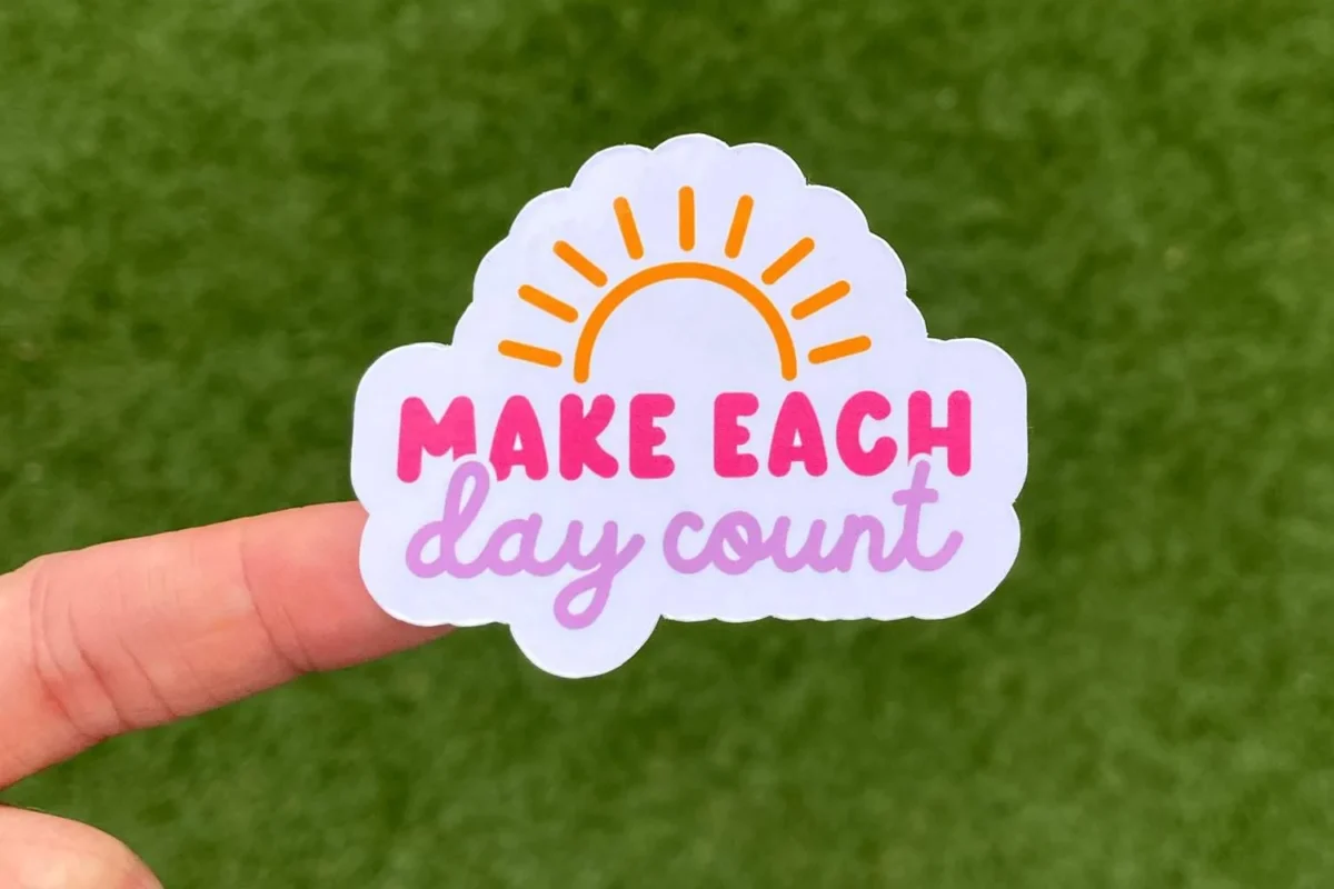 Hand holding a sticker that says “Make each day count” with a sun design