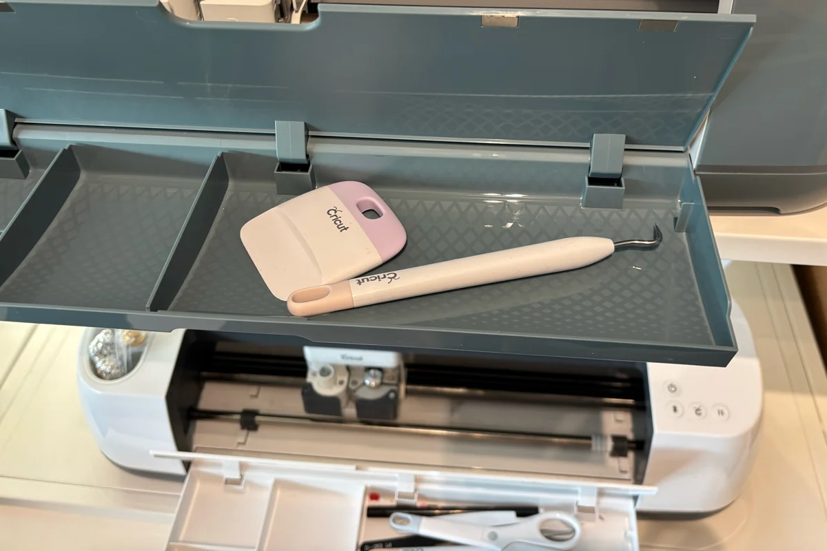 Cricut scraper and weeding tool stored inside the open accessory tray of a Cricut Maker machine, showing the built-in tool storage compartments.