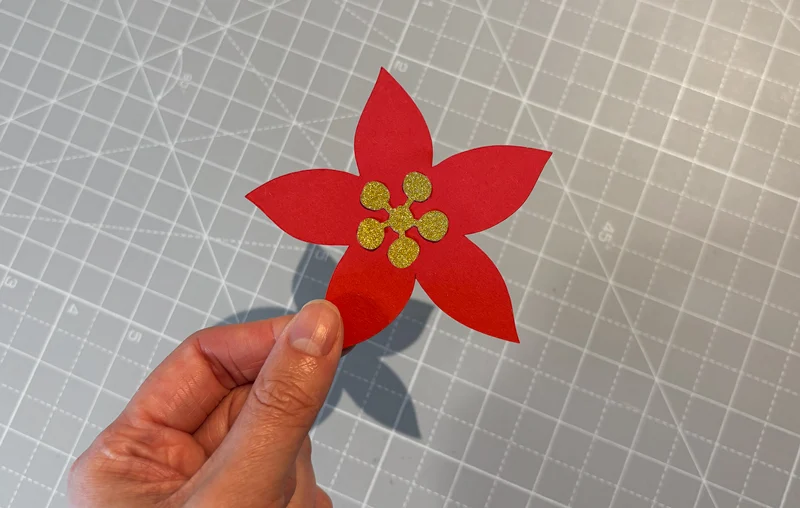 Hand holding a red paper poinsettia layer with gold glitter center pieces on a cutting mat