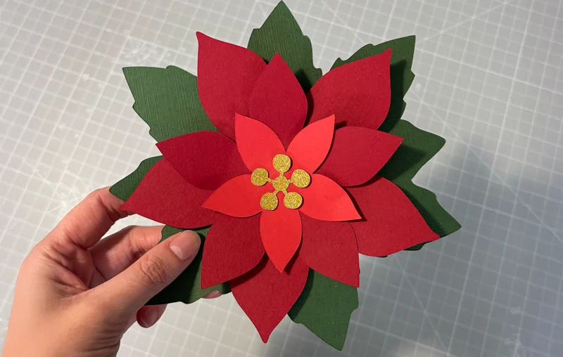 Hand holding a layered paper poinsettia flower with red petals, green leaves, and yellow center stamen
