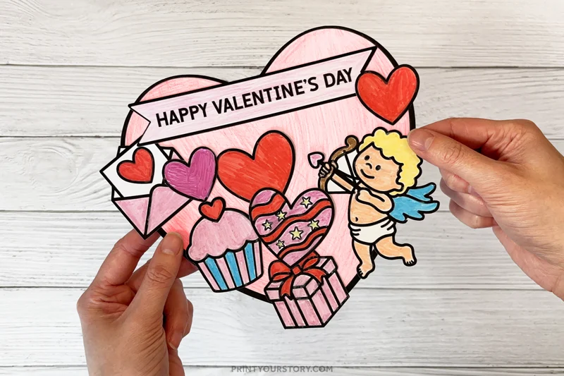 Handmade Valentine’s Day cut-and-paste coloring card with hearts, cupcake, gift, and cupid held in hands