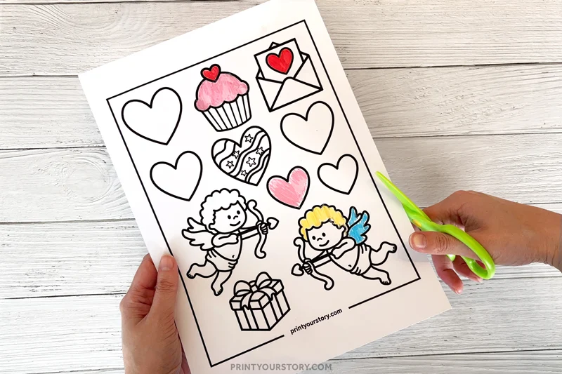 Child cutting a Valentine’s Day coloring card worksheet with hearts, cupids, and a gift