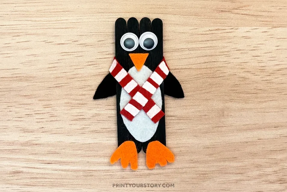 Penguin popsicle stick craft with googly eyes, orange felt beak and feet, and a red and white striped scarf