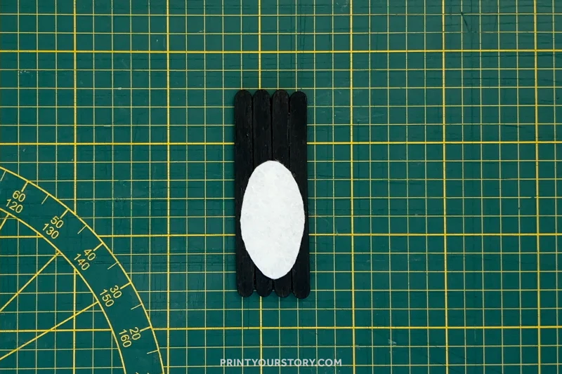 Black popsicle sticks with a white oval belly for a penguin craft on a cutting mat