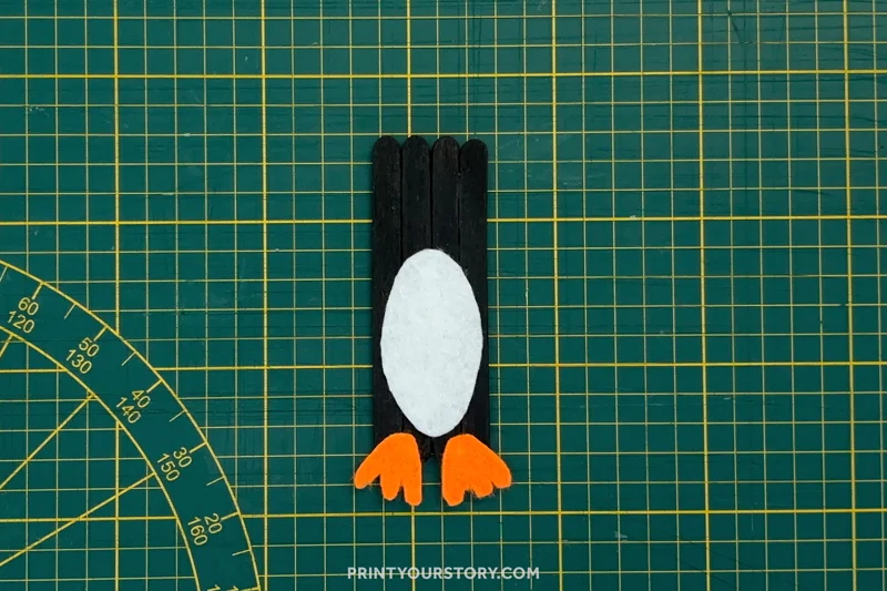 Black popsicle stick penguin craft with a white belly and orange feet on a cutting mat