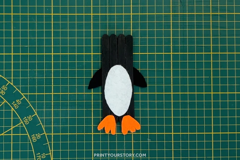 Popsicle stick penguin craft with white belly, black flippers, and orange feet on a cutting mat