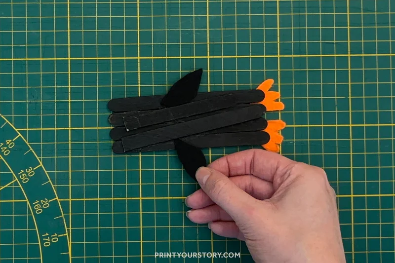 Hand attaching a black paper flipper to a popsicle stick penguin craft on a cutting mat