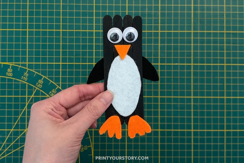 Hand holding a penguin popsicle stick craft with googly eyes, orange beak, felt belly, and feet