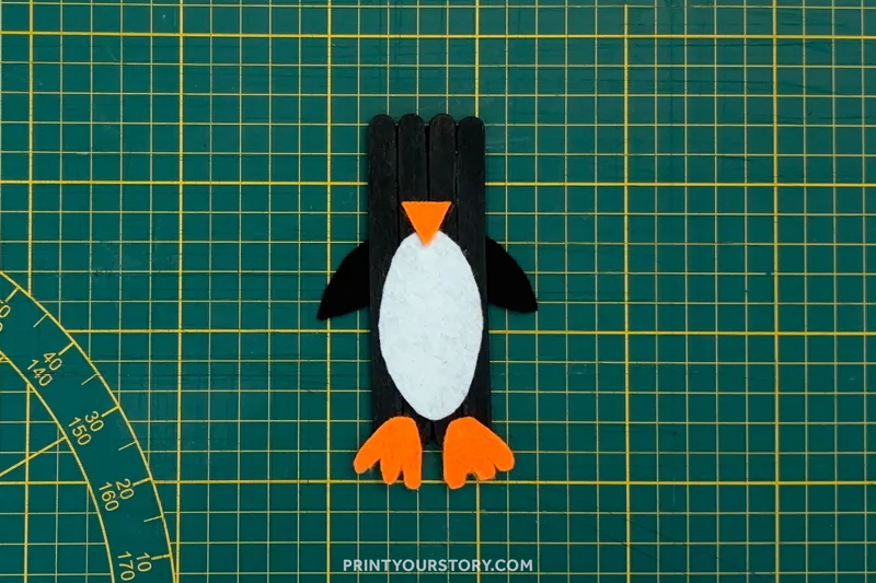 Penguin popsicle stick craft with white belly, orange beak, and feet on a green cutting mat
