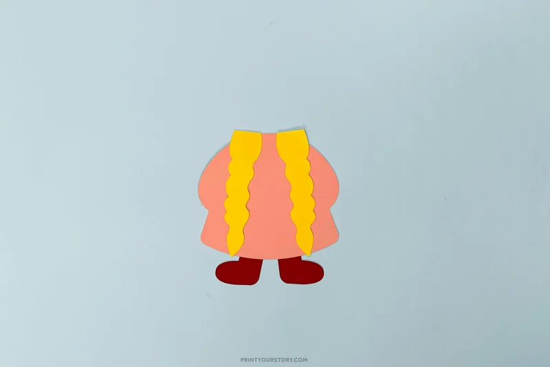 Paper Valentine gnome craft with a pink body, yellow braids, and red boots on blue paper