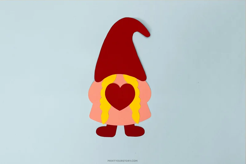 Valentine gnome craft with red hat, heart, and yellow braids on blue paper
