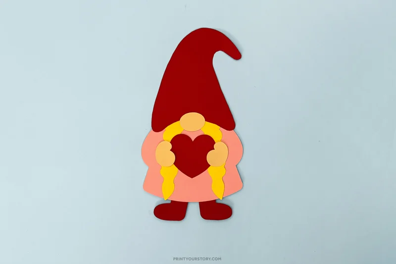 Paper Valentine gnome craft with a red hat, braids, and heart on a light blue background