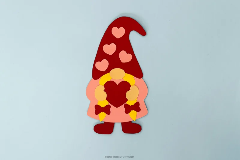 Paper Valentine gnome craft with red hat, heart decorations, and heart-shaped beard on blue background