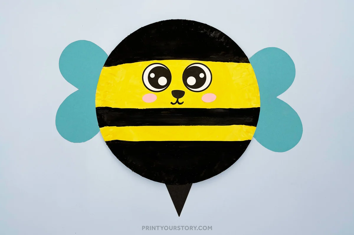 Paper plate bumble bee craft with black and yellow stripes, blue wings, kawaii eyes, cheeks, smile, and a black stinger