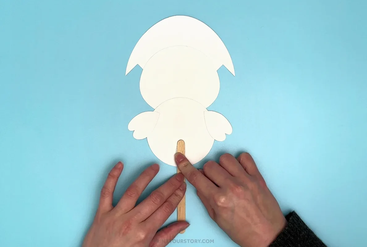 Hands attaching a popsicle stick slider to the back of a paper hatching chick craft