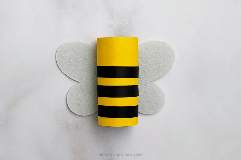 Toilet paper roll wrapped in yellow paper with black stripes for a bee craft. White felt wings. Marble gray background.