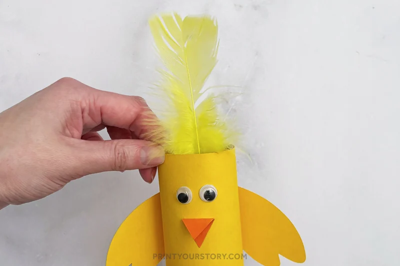 Hand placing yellow feathers inside a toilet paper roll chick craft with googly eyes and orange beak