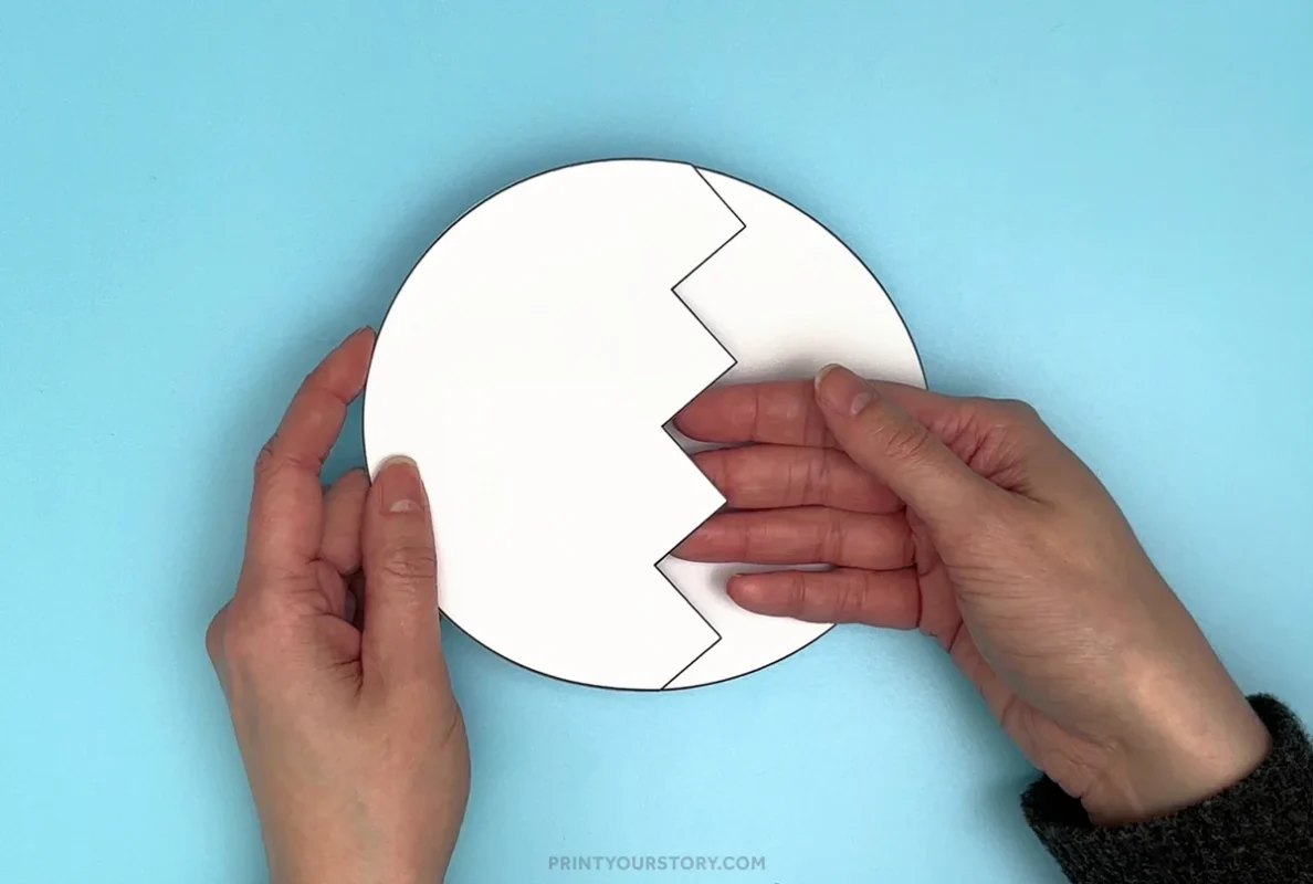 Hands holding a paper egg craft with a zigzag crack cutout on a blue background