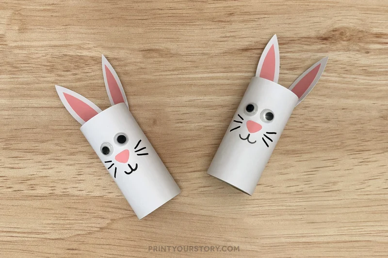 Two white toilet paper roll bunny crafts with paper ears and googly eyes on a wooden surface