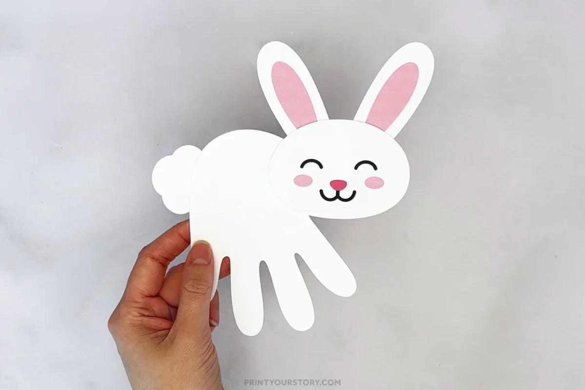 Hand holding a white paper handprint bunny craft with pink ears and cheeks