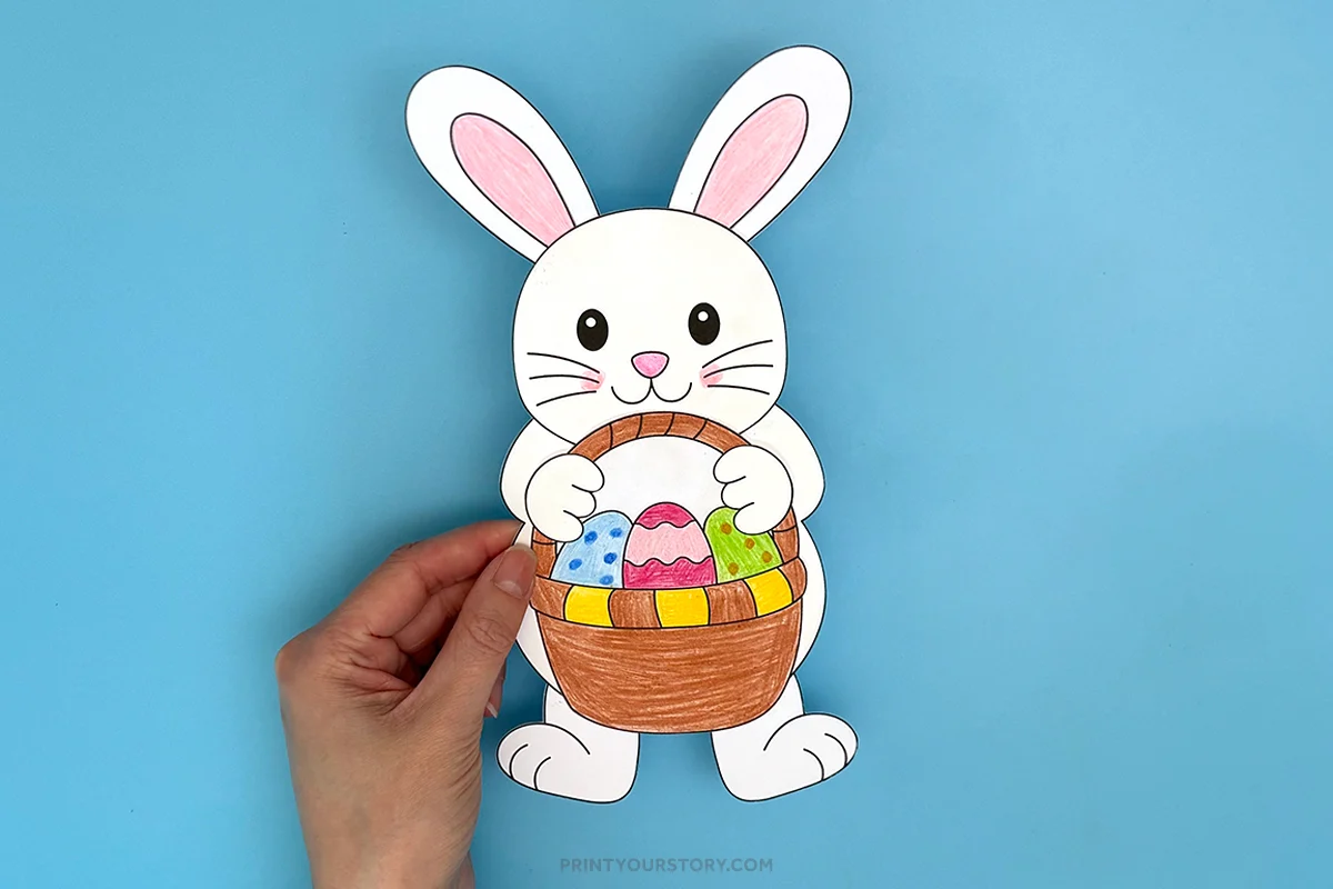 Hand holding a paper Easter bunny craft with a basket of decorated eggs on a blue background