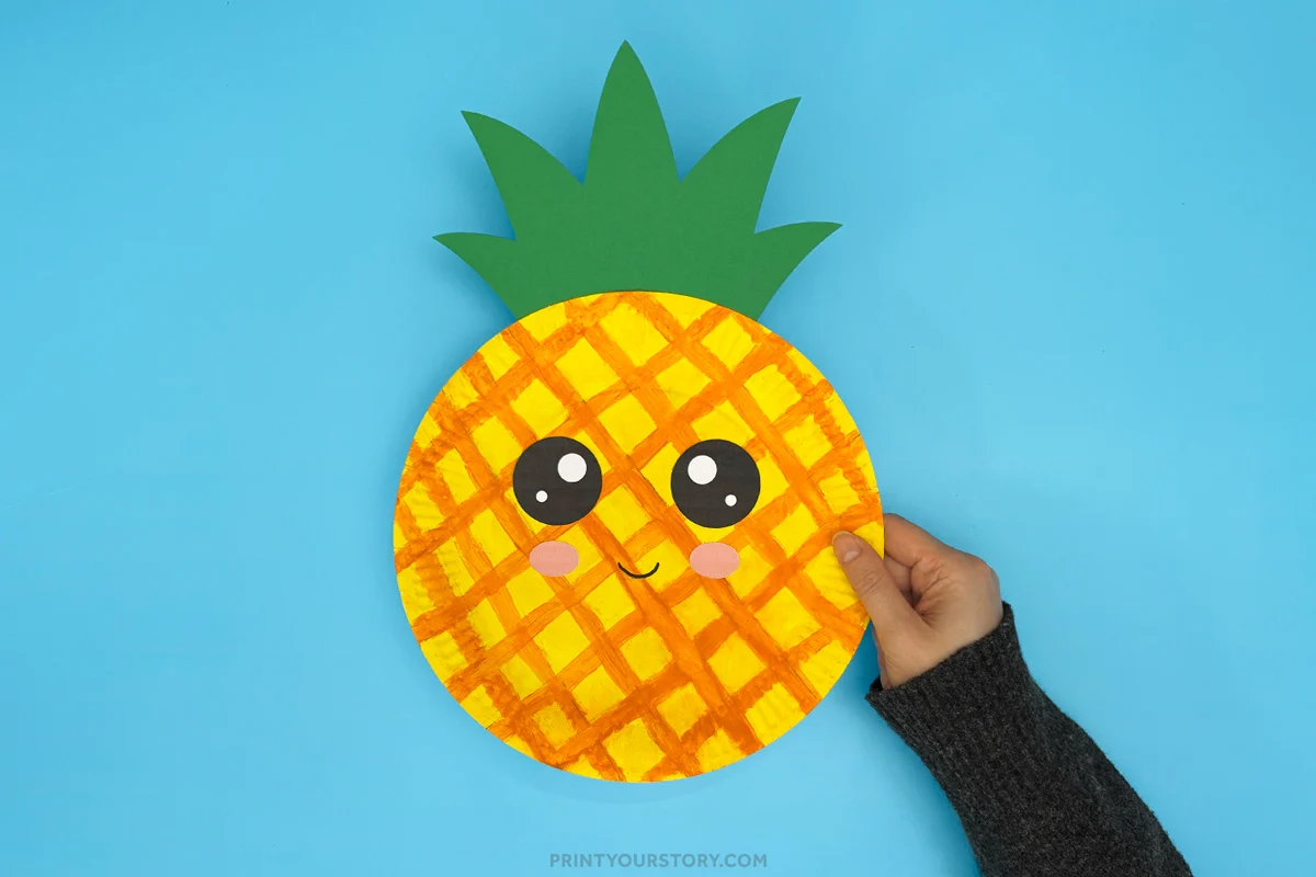 Hand holding a finished pineapple paper plate craft with painted pineapple texture, green paper leaves, large cartoon eyes, pink cheeks, and a smiling face on a blue background.