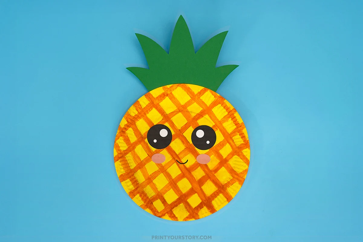 Finished pineapple paper plate craft with painted pineapple pattern, green cardstock leaves, large cartoon eyes, rosy cheeks, and a smiling face on a blue background.