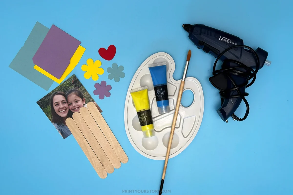 Materials for a popsicle stick Mother’s Day photo frame craft on a blue background