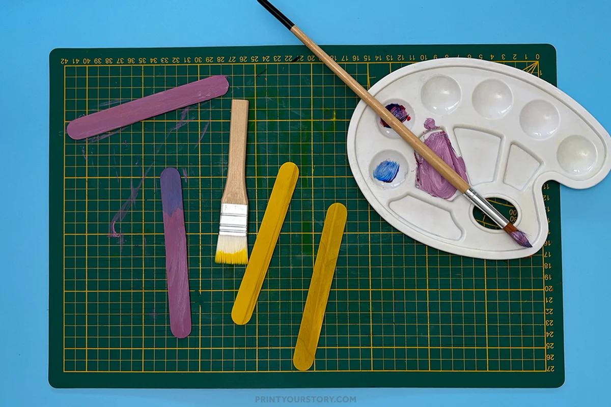 Painted popsicle sticks, brushes, and paint palette on a cutting mat for a Mother’s Day photo frame craft