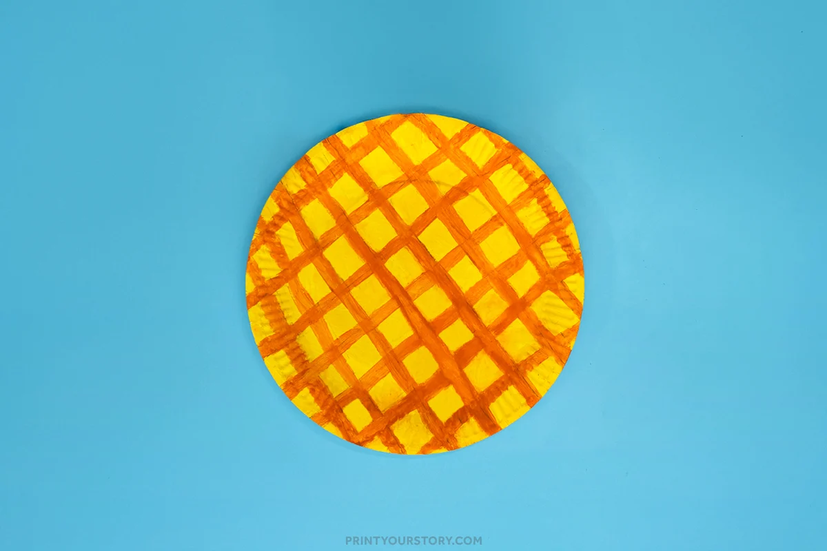 Painted paper plate pineapple craft base featuring yellow paint and orange crisscross lines on a blue background, ready for the next assembly step.