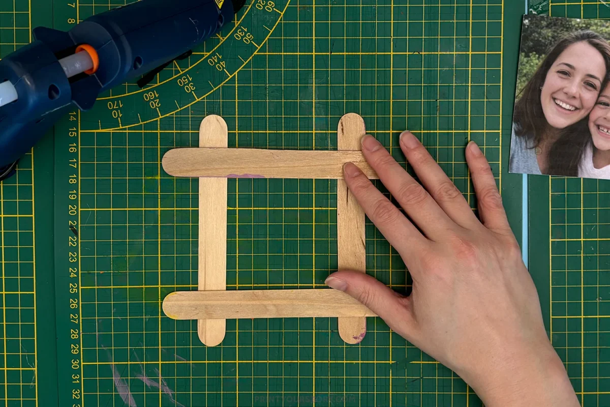Popsicle sticks arranged into a square photo frame on a cutting mat with a glue gun and photo nearby