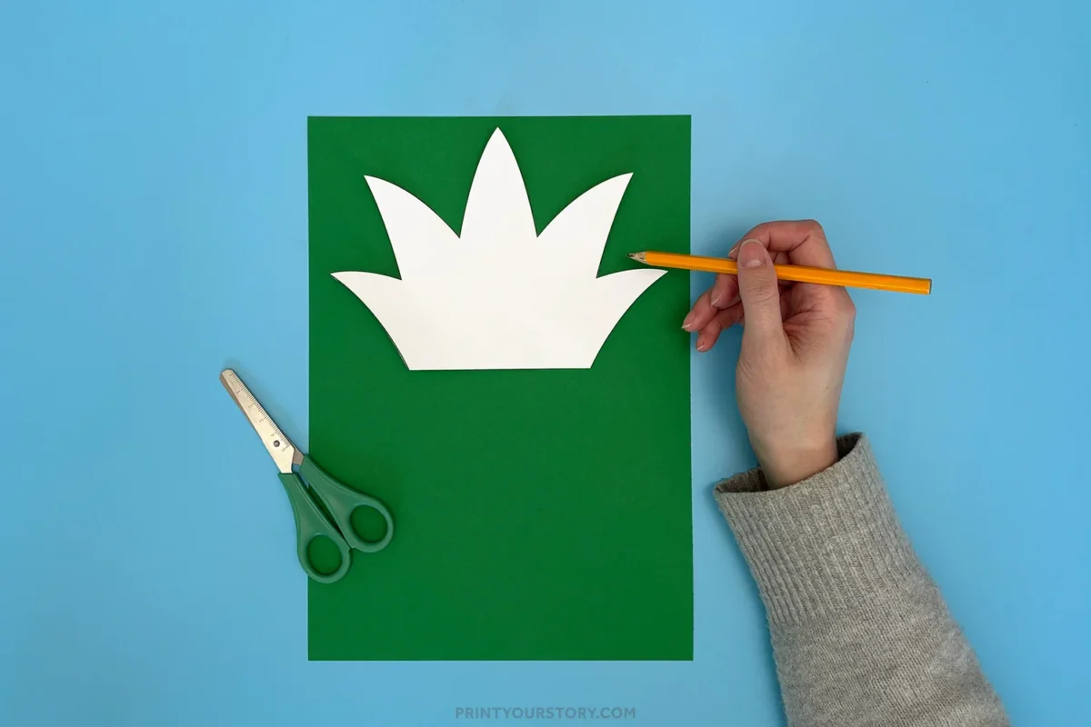 Hand tracing a printable pineapple leaf pattern onto green paper for a pineapple paper plate craft, with scissors nearby on a blue background.
