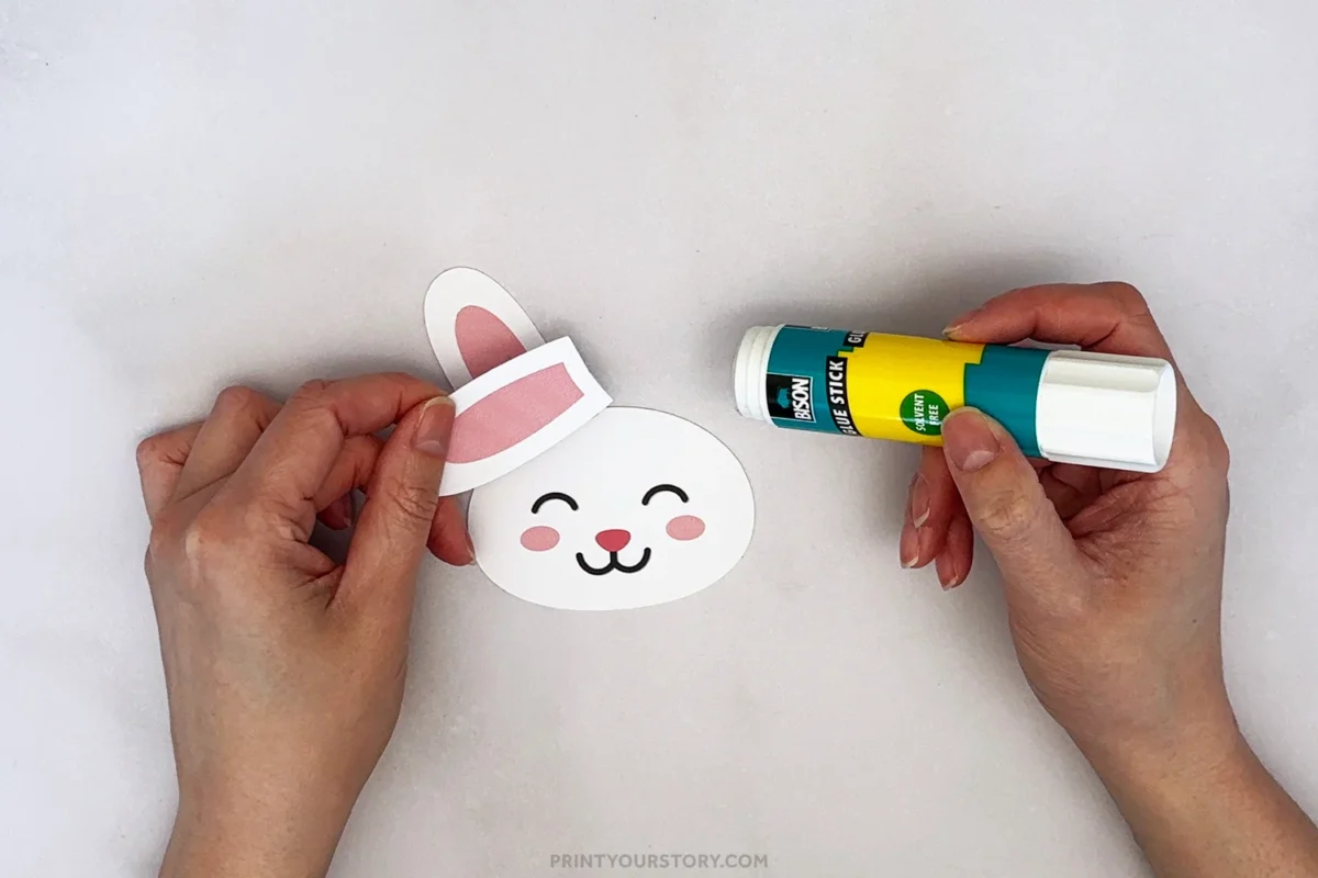 Hands holding a paper bunny face cutout and glue stick during a handprint bunny craft
