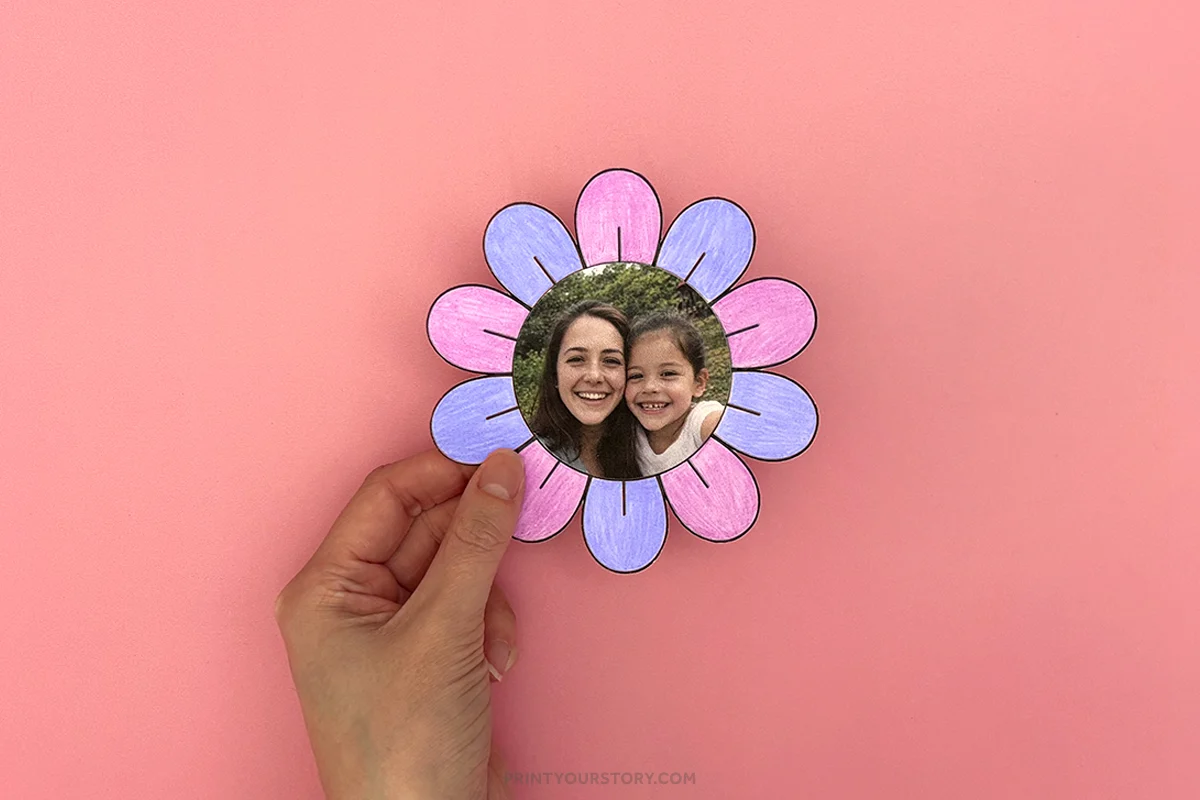 Hand holding a paper flower craft with pink and purple petals and a photo center on a pink background