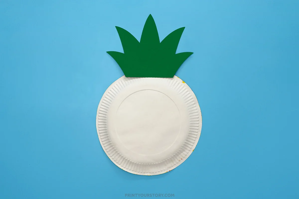 Back view of a pineapple paper plate craft showing a green cardstock leaf topper glued to the back of a paper plate on a blue background.