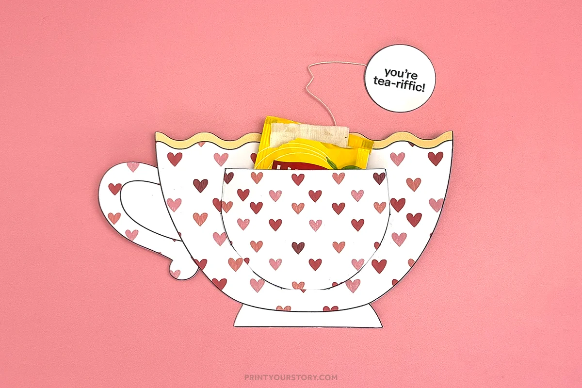 Heart-patterned paper teacup craft with tea bags and a “You’re tea-rific!” tag on a pink background