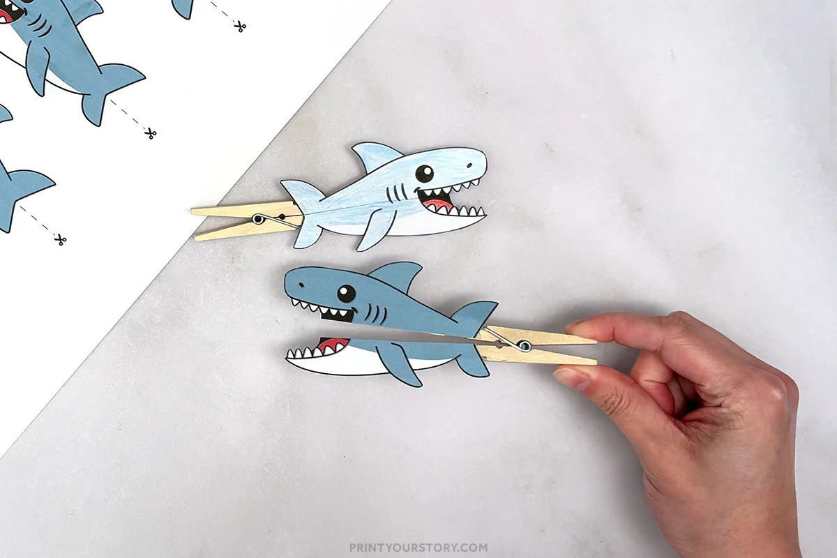 Printable shark clothespin puppet craft for kids with a completed colored shark puppet and a hand opening the shark's mouth