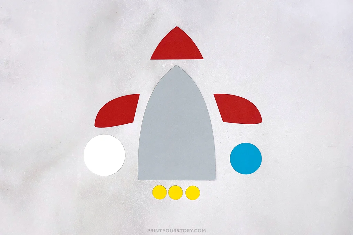 Paper pieces arranged to make a rocket handprint craft, with gray body, red fins, and colored circles