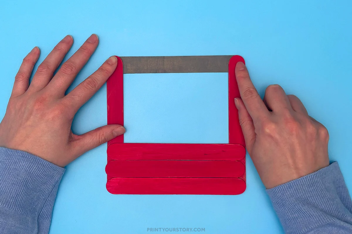 Hands arranging red popsicle sticks and a gray handle for a toolbox craft