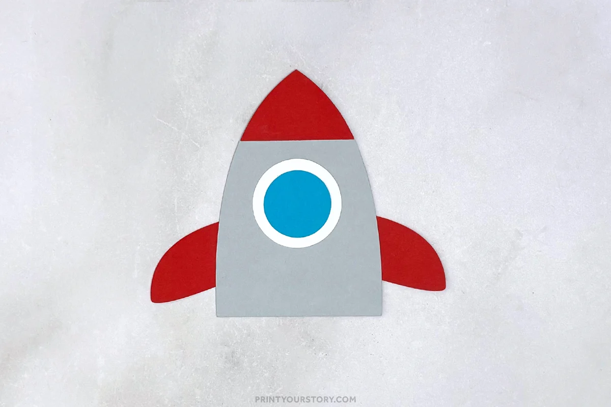 Paper rocket craft with red nose cone and fins, gray body, and blue window on a light background