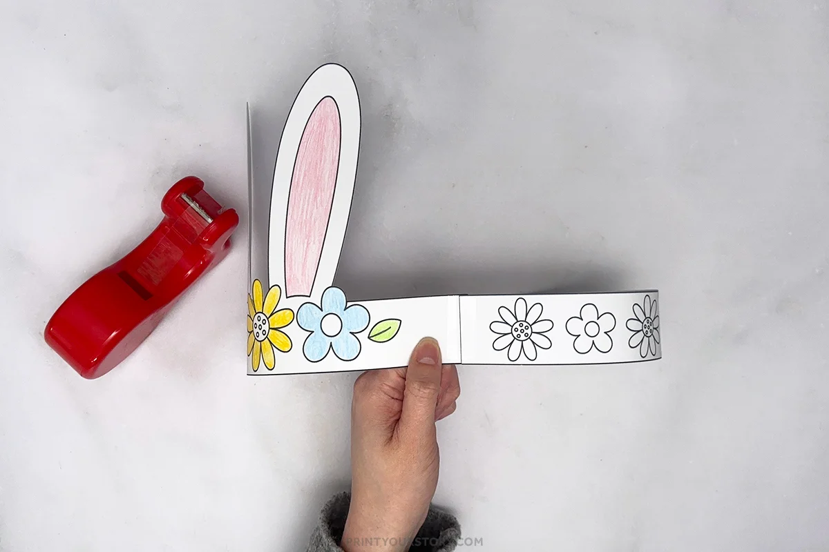 Hand holding a printable bunny headband with a pink ear and flower band, with a red stapler nearby