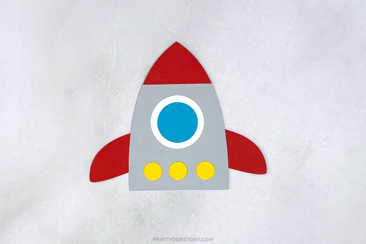 Paper rocket craft with red fins, gray body, blue window, and yellow circles on a light background