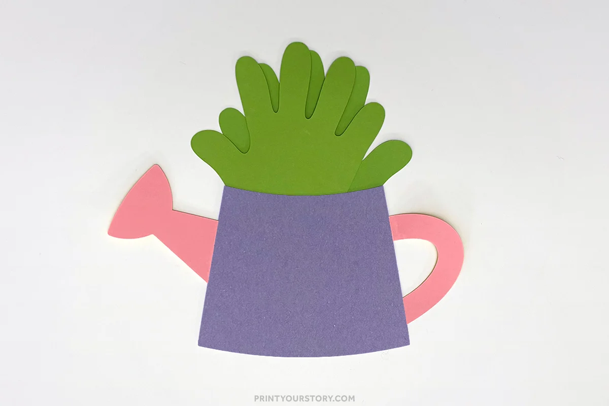 Paper watering can craft with green handprint leaves, purple body, and pink spout and handle