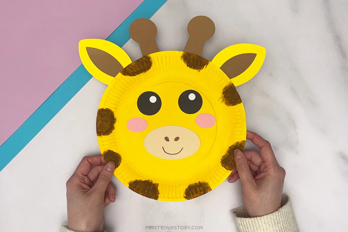 Hands holding finished giraffe paper plate craft with ears, horns, spots, and smiling face