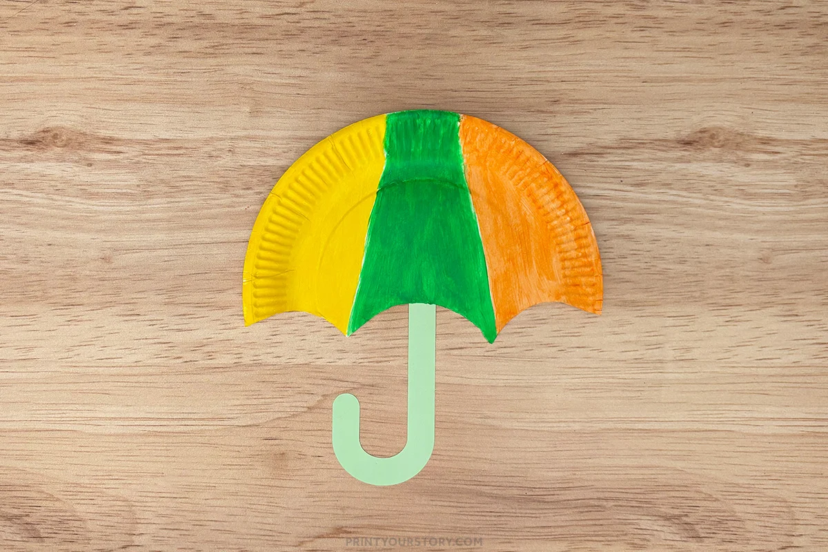 Colorful paper plate umbrella craft with a green J-shaped handle on a wooden surface