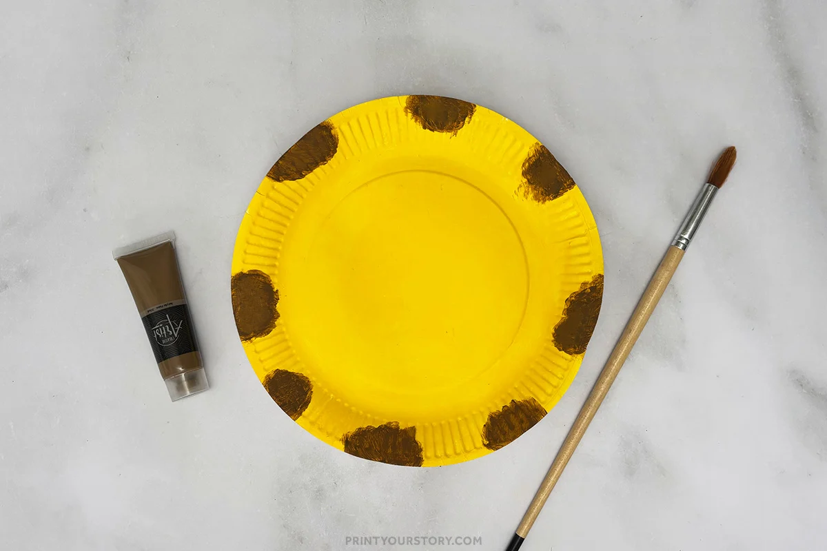 Yellow paper plate with brown giraffe spots painted around the rim, with paint and brush nearby